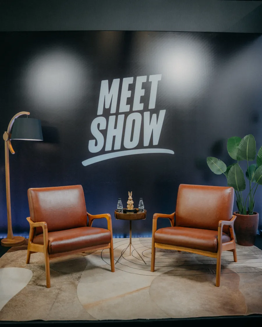Palco do Meet Show com duas poltronas vazias diante do painel com a marca